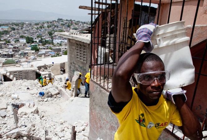 Haiti - Sáu tháng sau thảm họa Một người dân ở thủ đô Port-au- Prince đi nhặt nhạnh những gì còn sót lại trong một ngôi nhà đổ nát. Chiếc áo anh mặc, giống như của nhiều người khác, là hàng viện trợ của chính phủ Mỹ. Ảnh: Getty Images