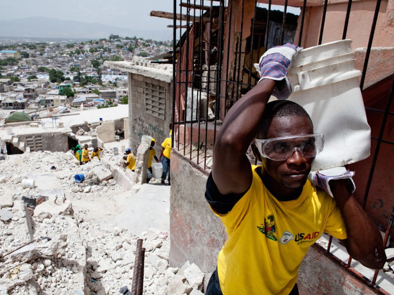 Một người dân ở thủ đô Port-au- Prince đi nhặt nhạnh những gì còn sót lại trong một ngôi nhà đổ nát. Chiếc áo anh mặc, giống như của nhiều người khác, là hàng viện trợ của chính phủ Mỹ. Ảnh: Getty Images