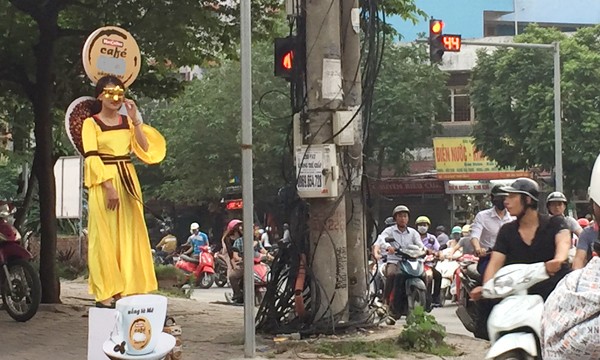 Cô gái trẻ mặc váy vàng, mặt đeo mặt nạ đứng vỉa hè gần nút giao thông Trung Kính- Vũ Phạm Hàmgây chú ý cho người đi đường