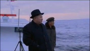 Ông Kim Jong-un đứng trên một chiếc tàu quân sự, mặc áo khoác, đầu đội mũ, quan sát vụ phóng tên lửa từ tàu ngầm. Ảnh cắt từ clip