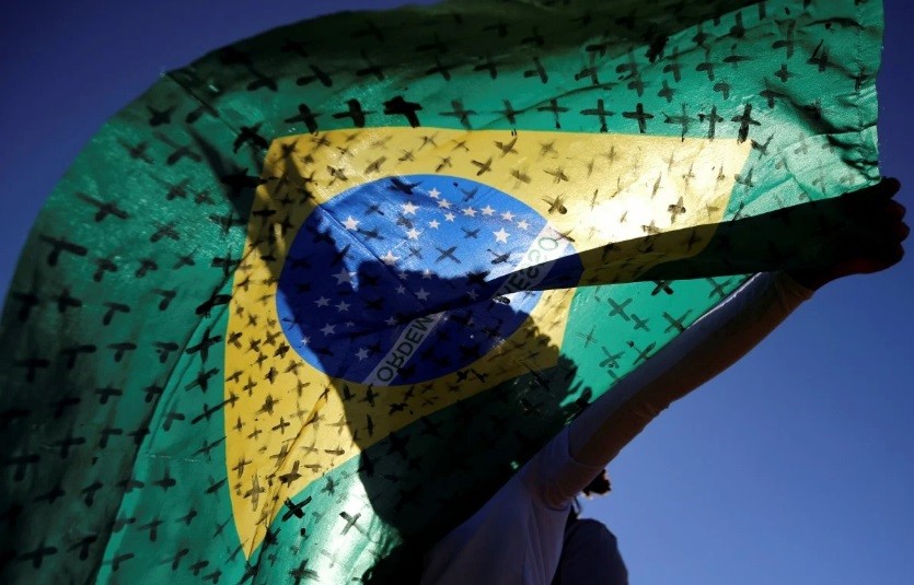 Một nhà hoạt động cầm quốc kì Brazil có hình thánh giá tượng trưng cho những người đã qua đời vì COVID-19. Ảnh: Reuters