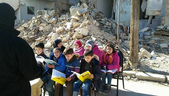 Cảnh các em nhỏ Syria chăm chú nhìn vào quyển sách ở một lớp học bên ngoài trời giữa cảnh đổ nát bao quanh.